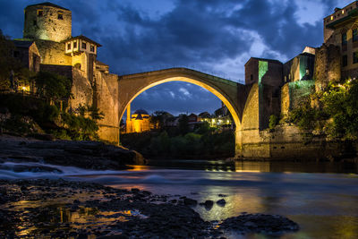 Arch bridge over river by buildings against sky at dusk