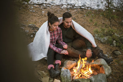 Couple wrapped in blanket while sitting by fire pit at campsite