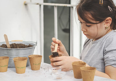 A caucasian girl enthusiastically fills the soil with a wooden spoon into a cardboard glass.