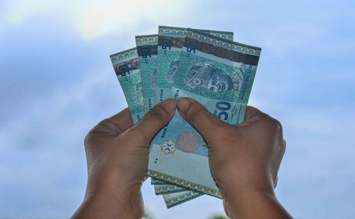 Cropped hands holding paper currency against sky
