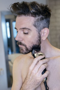 Shirtless man trimming beard at home