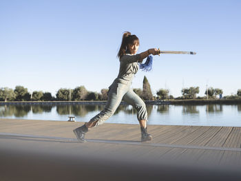 Female athlete practicing martial arts with sword by lake against clear sky