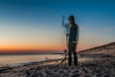 Double exposure of man standing by tripod on beach against sky during sunset