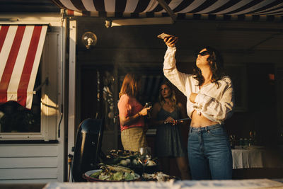 Woman photographing food on barbecue grill through smart phone while friends standing in background