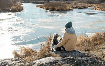 Young woman sitting on stone hugging dog looking at river fall or at beginning of winter, pet love