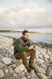 Man sitting on rock with camera at beach against sky