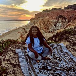 Portrait of smiling young woman sitting on rock at sunset
