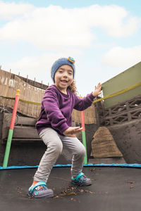 Full body of positive ethnic child in headband looking at camera with smile while jumping on trampoline and having fun in countryside