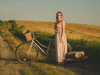 Portrait of woman with bicycle standing on grassy field against sky