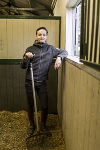 Full length portrait of confident young man with pitchfork in horse stable