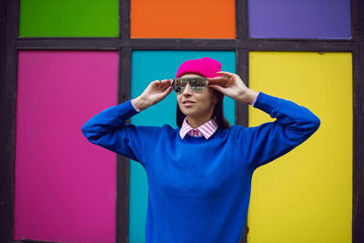 Fashionable brunette woman in a pink warm hat and sweater and sunglasses stands against background