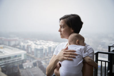 Mother carrying son looking away while standing in balcony during foggy weather