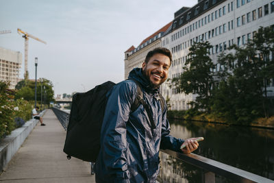 Portrait of smiling male delivery person holding smart phone while standing near railing on footpath