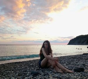 Young woman sitting on rock at beach against sky during sunset
