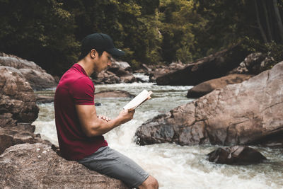 Side view of man reading book while sitting on rock by stream