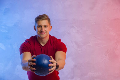 Portrait of smiling young man holding exercise ball against wall