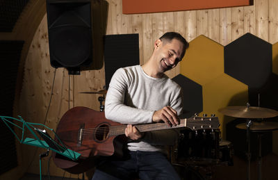 Smiling white man tuning guitar, playing, holding musical instrument in hands, sitting on chair in