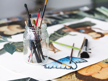 Glass jar with assorted paintbrushes placed on various creative paintings on table in workshop