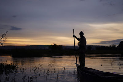 Man standing on boat amidst lake against sky during sunset