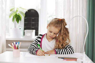 A charming girl sits at a table and draws a rainbow in an album with colored pencils. homework