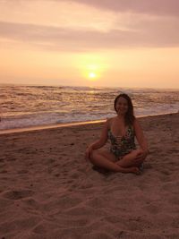 Young woman sitting on shore at beach against sky during sunset