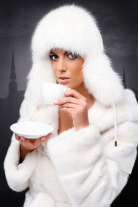 Portrait of young woman in fur coat holding cup and saucer against wall