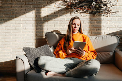 Full length of woman using digital tablet sitting on sofa at home