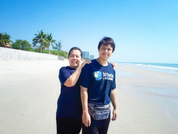Portrait of smiling mature couple standing at beach against clear blue sky during sunny day