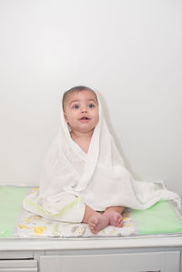 Thoughtful cute baby boy wrapped in towel while sitting on bed against wall