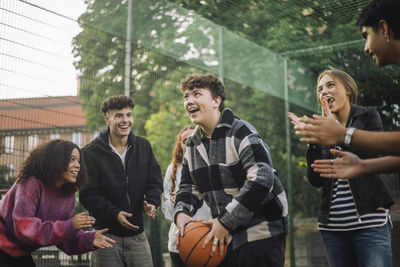 Happy friends cheering teenage boy preparing to take shot while playing basketball at court