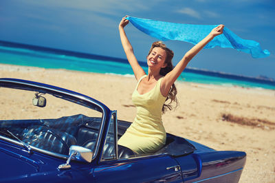 Smiling woman holding aloft scarf while sitting in car by sea against sky