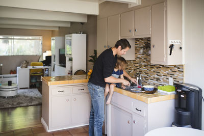 Father washing kitchen utensil while daughter sitting on kitchen counter