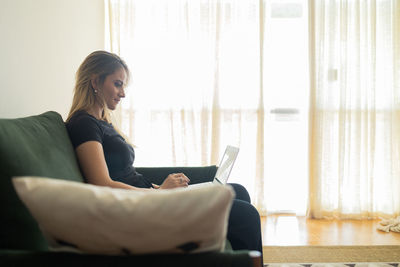 Woman working at home from couch with laptop on her lap. home office concept. gray notebook