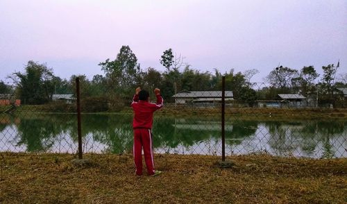 Rear view of boy looking through fence by lake against sky