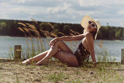 A woman in a swimsuit, hat and sunglasses sunbathes in summer on the riverbank among the grass