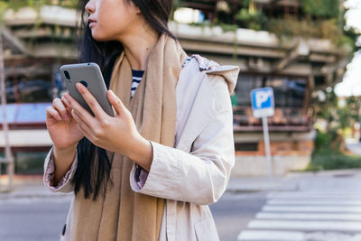 Cropped unrecognizable asian female standing on the city street browsing on mobile phone looking away in sunny day