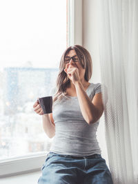 Woman with eyeglasses and curly hair sits on windowsill. sleepy woman yawns. morning cup of coffee.