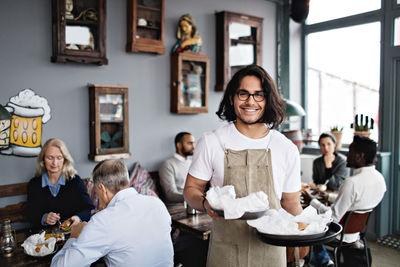 Portrait of smiling young waiter serving food while standing against customers at restaurant