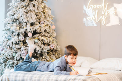 Boy doing homework lying on the sofa, against the backdrop of a christmas tree, in his room