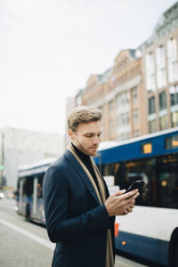 Confident entrepreneur surfing net through smart phone while standing in city