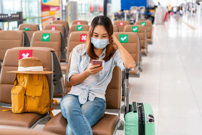 Woman wearing mask using mobile phone while sitting at airport