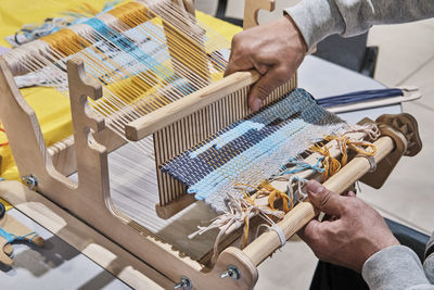 Hands of senior man weaving small rug with pattern on manual table loom, at masterclass on weaving.