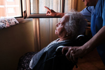 Side view of copped unrecognizable caring nurse in uniform pointing away while standing near sick elderly female in wheelchair near window in light room