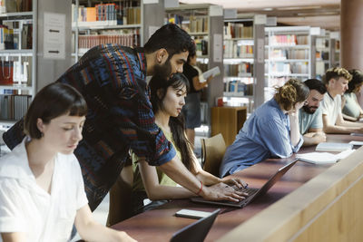 Man assisting female friend using laptop at desk in college library