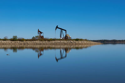 Oil pump jacks on a rocky peninsula with their reflections in the glassy water of a lake.