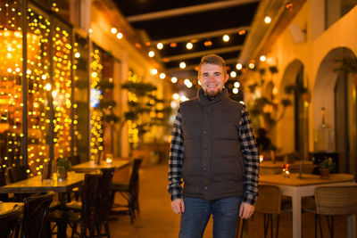 Portrait of young woman standing against illuminated restaurant