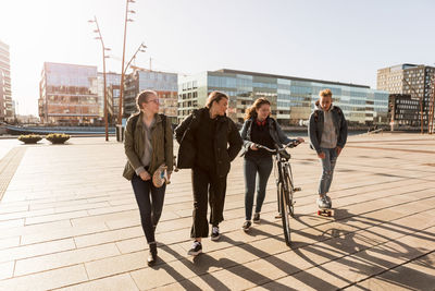 Teenage girl walking with bicycle and friends while boy riding bicycle at city