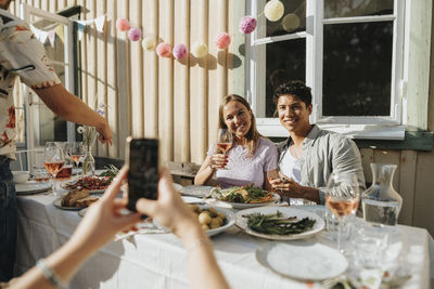 Woman clicking picture of male and female friends holding wineglass during dinner party at cafe