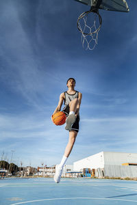 From below of anonymous sportive male passing the ball under his legs into basketball hoop while playing game against blue sky on sunny summer day