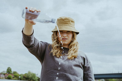 Low angle portrait of young woman pouring water from bottle against sky